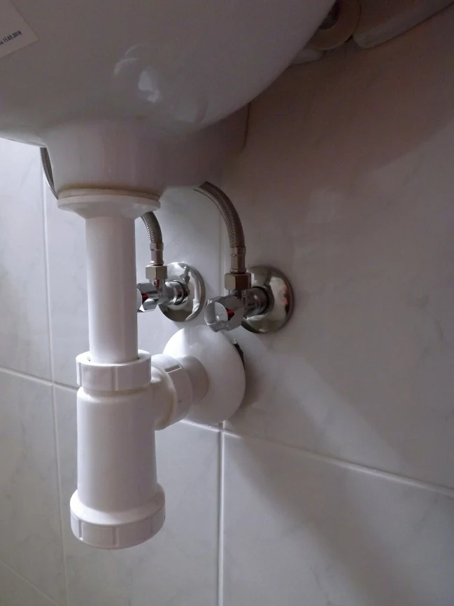 Bílá odpadní trubka s vodovodními ventily pod umyvadlem.