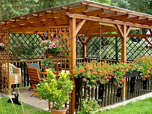 Dřevěná zahradní pergola s posezením a kvetoucími rostlinami.