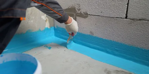 Pracovník nanáší modrou hydroizolační barvu na roh koupelny.