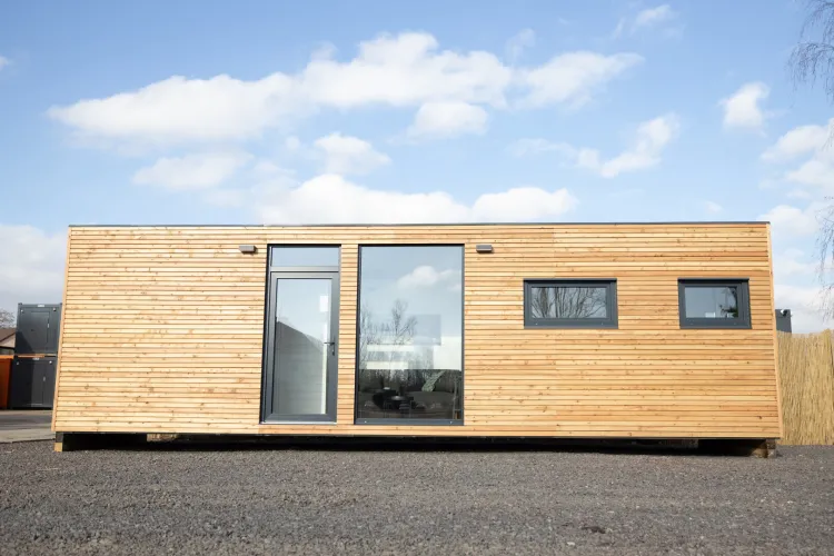 Moderní dřevěný kontejnerový dům s velkými okny na pozadí modré oblohy.