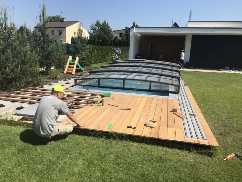 Montáž dřevěné terasy kolem bazénu s pracovníkem na trávníku.
