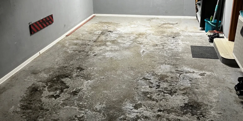 Poškozená betonová podlaha v garáži s plísní a trhlinami.