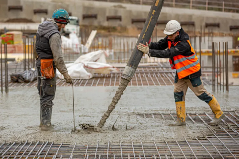 Dva dělníci nalévají beton na výztužnou mřížku ve stavbě.