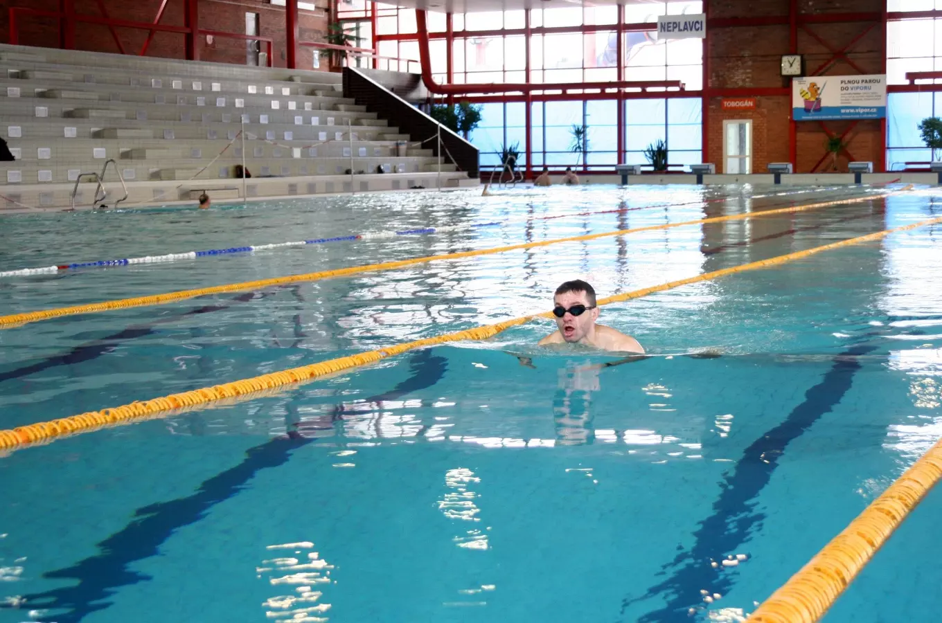 Muž plave na zádech v krytém bazénu s tribunou.