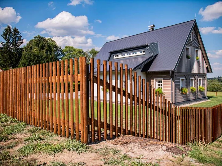 Dřevěný plot z kulatin obklopující dům na pozadí modrého nebe.