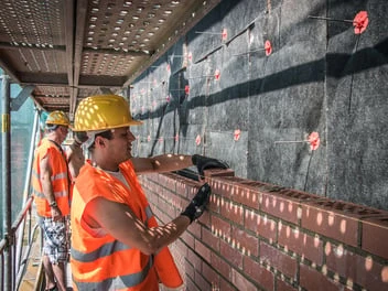 Stavebníci montují větranou fasádu z cihel pod střešním přesahem.