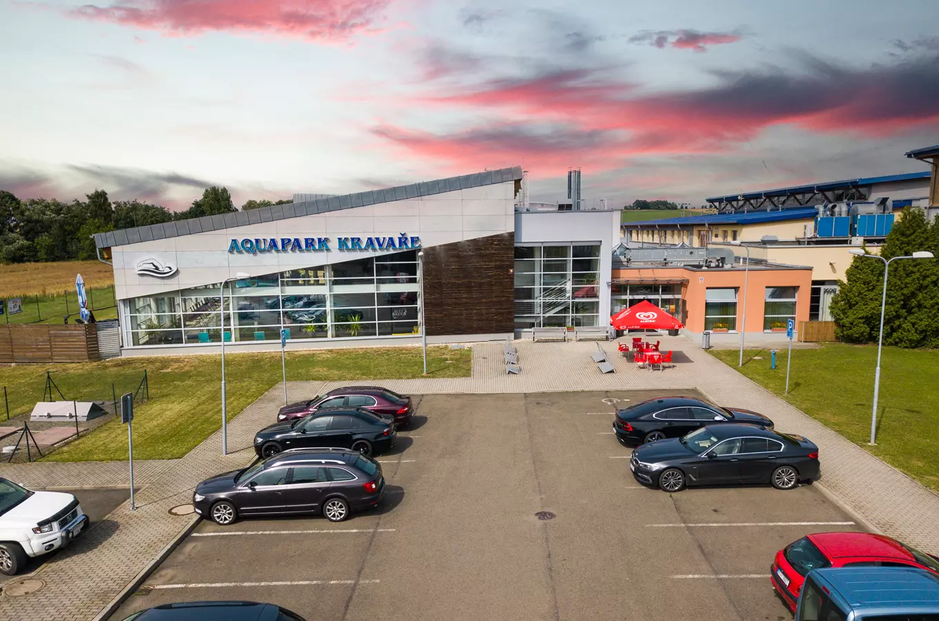 Vnější pohled na Aquapark Kravaře s parkovištěm aut pod barevnou oblohou.