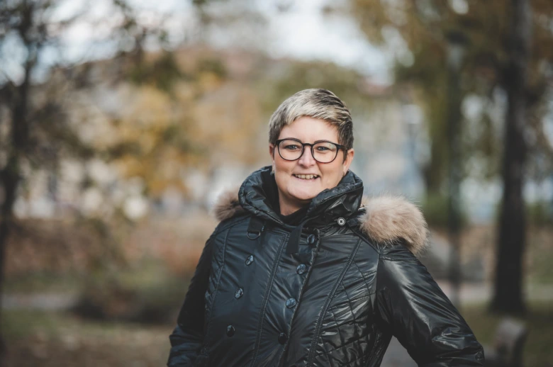 Žena v zimní bundě stojí venku na pozadí stromů.