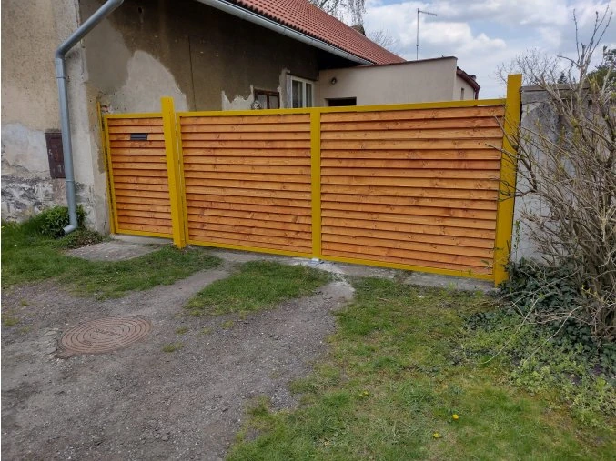 Dvoukřídlá dřevěná vjezdová brána s integrovaným plotem a žlutými rámy.