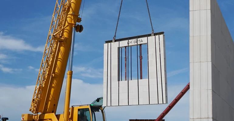 Žlutý kran zvedá prefabrikovaný betonový stěnový prvek při stavbě rodinného domu.