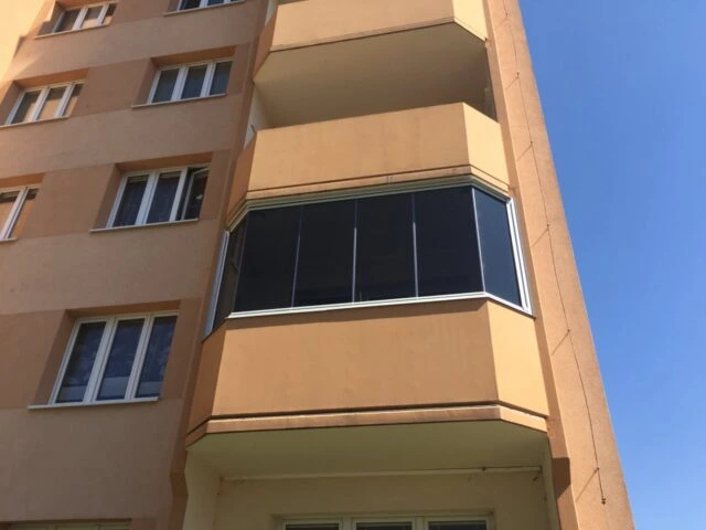 Zasklený balkon na fasádě bytového domu pod modrou oblohou.