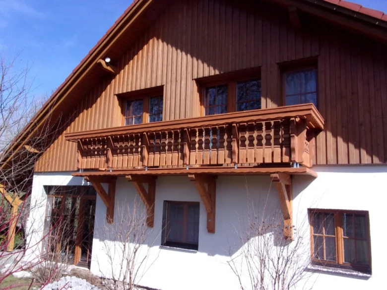 Dřevěný balkon na fasádě domu s bílými zdmi.