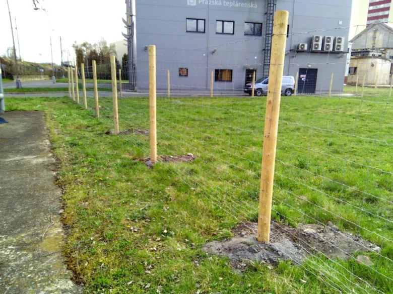 Řada dřevěných sloupků s drátovou plotem na trávníku u budovy.