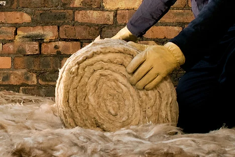 Pracovník s rukavicemi manipuluje se rolí minerální vlny před cihlovou zdí.