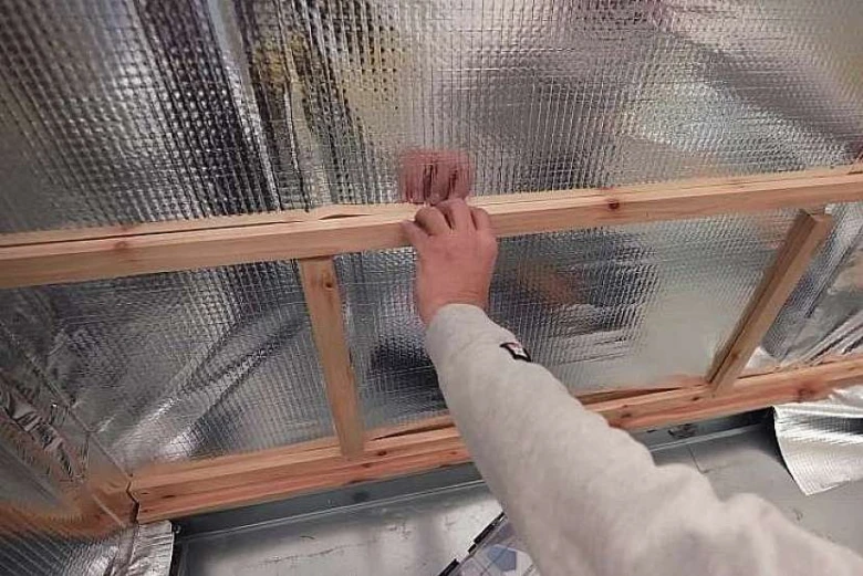 Ruční instalace dřevěného rámu nad izolačním fóliem při stavbě.