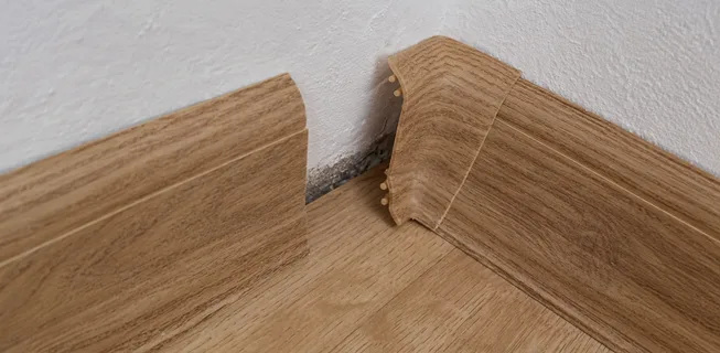 Poškozený roh vinylové podlahy u zdi s viditelným odpadem.