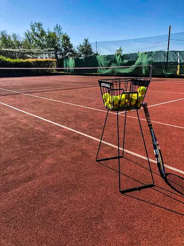 Tenisový dvorec s košíkem míčů a raketou na slunci.