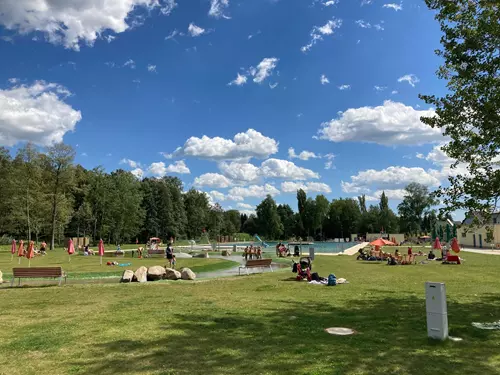 Venkovní bazén v Hlinsku s lidmi odpočívající na trávníku pod modrou oblohou.